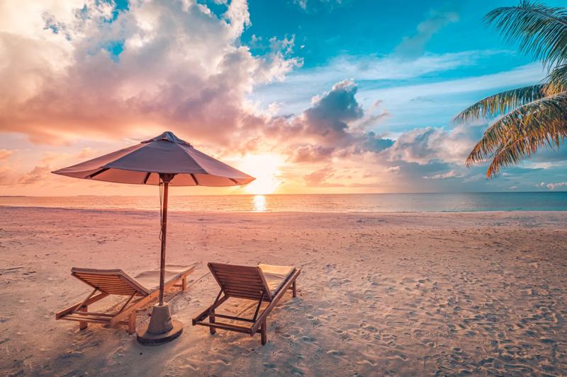 Beautiful tropical sunset scenery, two sun beds, loungers, umbrella under palm tree. White sand, sea view with horizon, colorful twilight sky, calmness and relaxation. Inspirational beach resort hotel