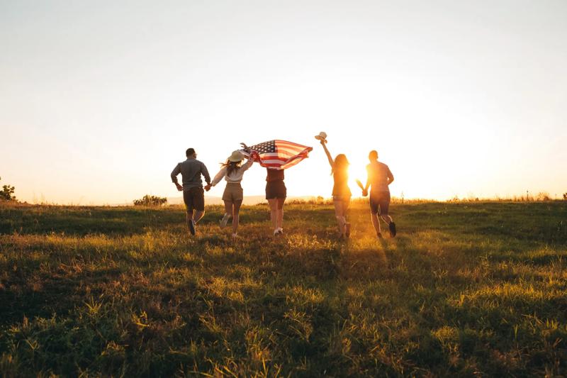 Group of young caucasian handsome people spreading wide american flag at sunset.