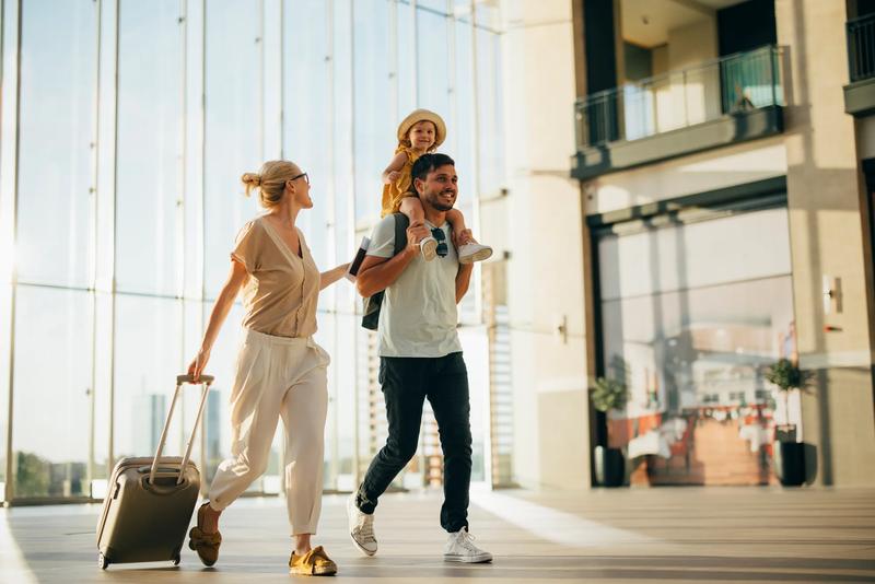 Cheerful husband carrying his cute daughter on shoulders while walking with his beautiful wife and luggage at the airport