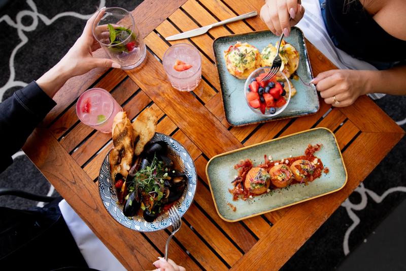 A view looking down onto a meal for two including a shrimp and eggs Benedict with a fruit cup, a bowl of mussels and an appetizer of hushpuppies